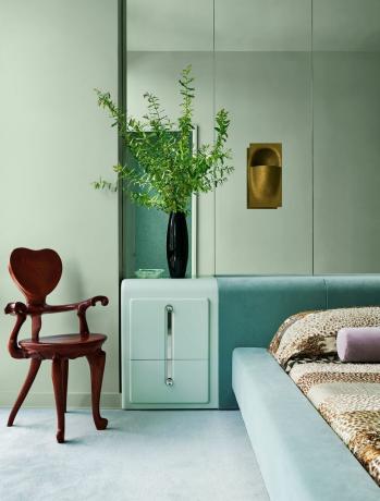 et soveværelse har mintgrønne vægge, en spejlvæg bagved en seng betrukket med et lyst aqua fløjl og et lysegrønt natbord, bronzelampet, en egetræslænestol med hjerteformet ryg, mintgrønt tæppe