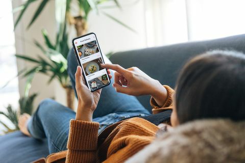 over skulderen visning af en ung kvinde, der vælger mad fra menuen i mobilappen, mens hun ligger i sofaen derhjemme