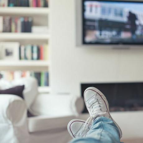 Billede af jeans og sneakers, fødderne op på sofaen, med tv, pejs, sofa og bogreol i baggrunden.